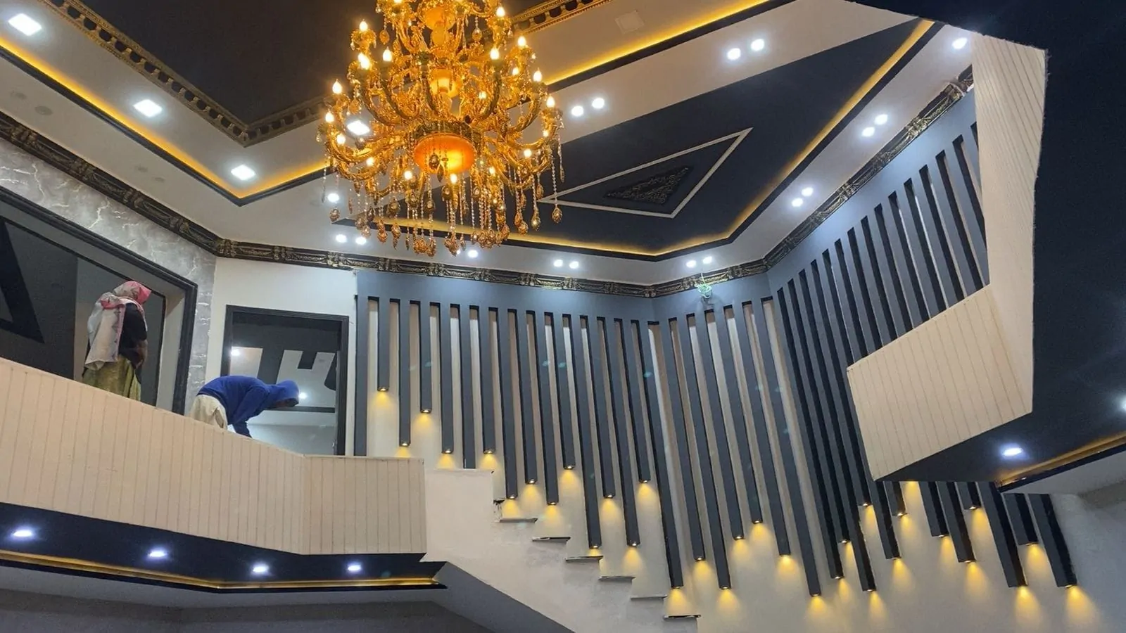 Double-height staircase hall with backlit wall battens and a gold chandelier